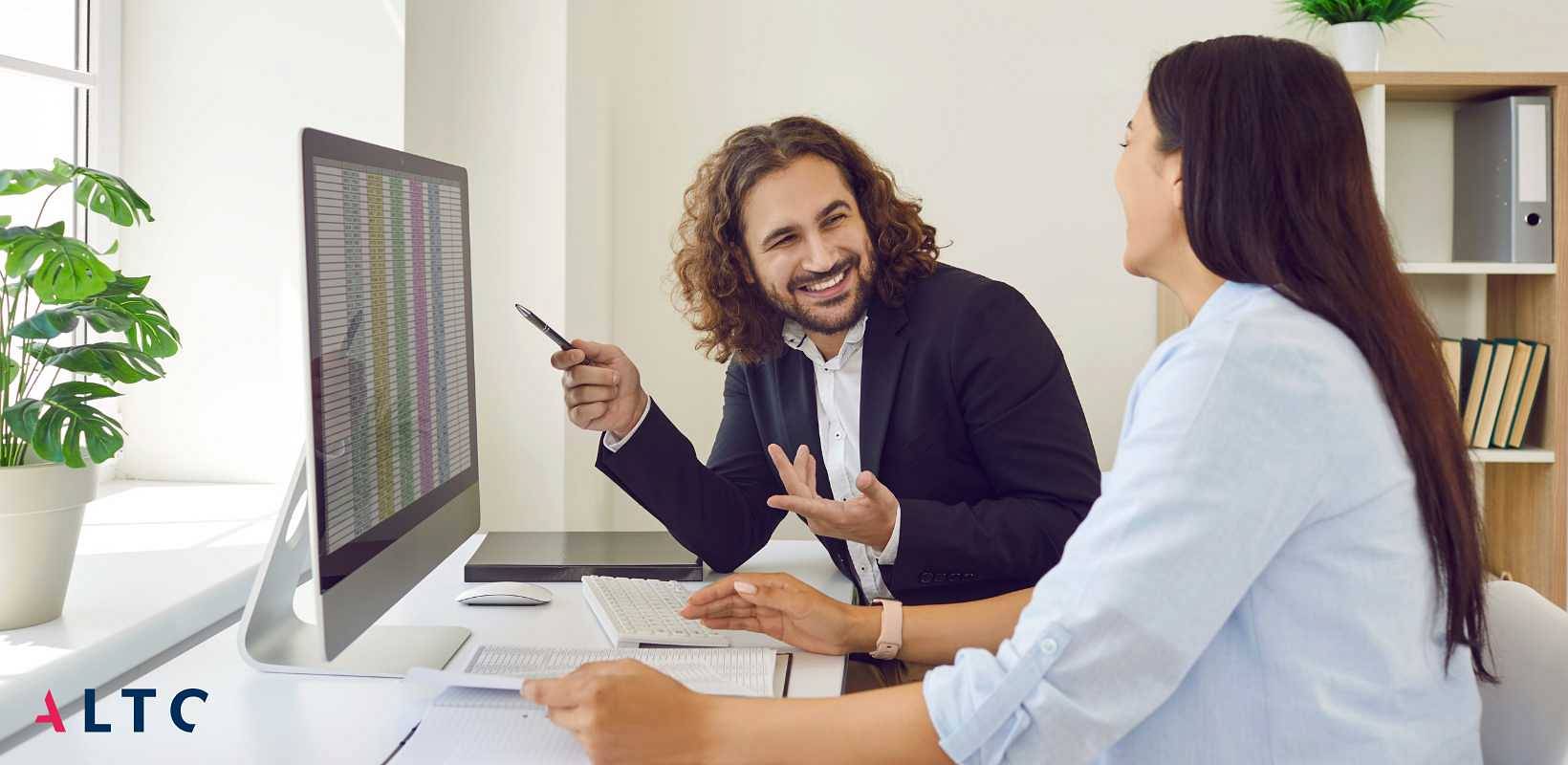 Uśmiechnięty mężczyzna w formalnym stroju omawia coś z kobietą w biurze, wskazując długopisem na ekran monitora, na którym widoczne są dane w formie tabeli.jpg